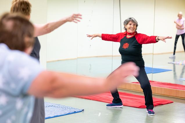 Eine ältere Frau in Sportkleidung führt eine Dehnübung in einem Fitnessraum mit Spiegeln aus.